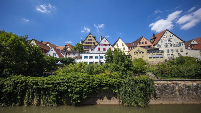 Bunte Fachwerkhäuser auf einem Hügel über einem Fluss, umgeben von Bäumen, unter blauem Himmel., © Stuttgart Marketing GmbH Achim Mende