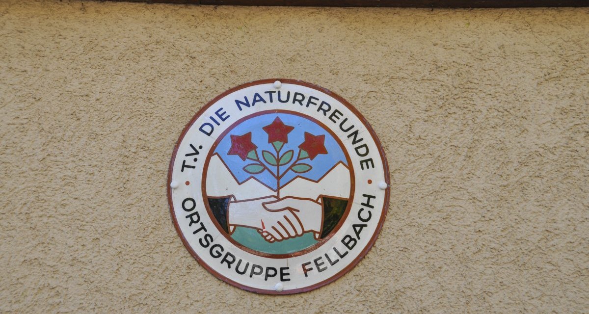 Rundes Logo der Naturfreunde Ortsgruppe Fellbach auf einer beigen Wand. Es zeigt zwei Hände, die sich schütteln, und drei rote Blumen vor Bergkulisse. Rundes Logo der Naturfreunde Ortsgruppe Fellbach auf einer beigen Wand. Es zeigt zwei Hände, die sich schütteln, und drei rote Blumen vor Bergkulisse.
