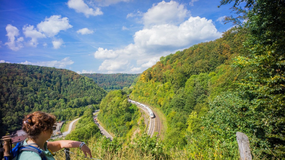 Ein Wanderer blickt vom Mühltalfelsen auf die kurvenreiche Geislinger Steige, wo ein Zug durch die grüne Landschaft fährt., © Landkreis Göppingen