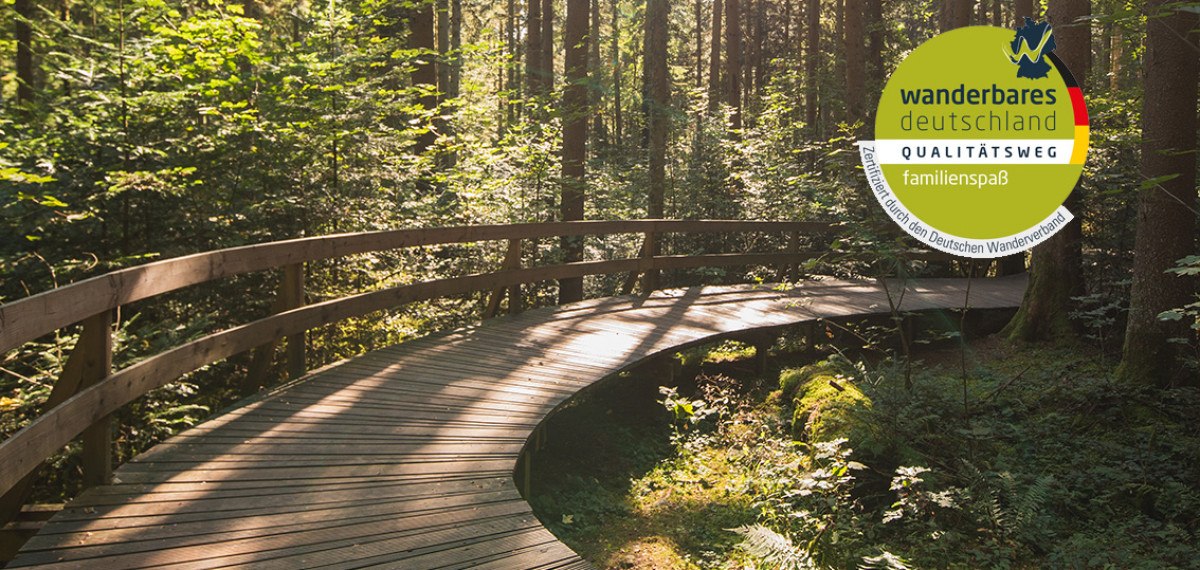Ein geschwungener Holzweg führt durch einen sonnendurchfluteten Wald. Ein Siegel von 'Wanderbares Deutschland' ist sichtbar., © Touristik und Marketing GmbH Schwäbisch Gmünd Bebop Media