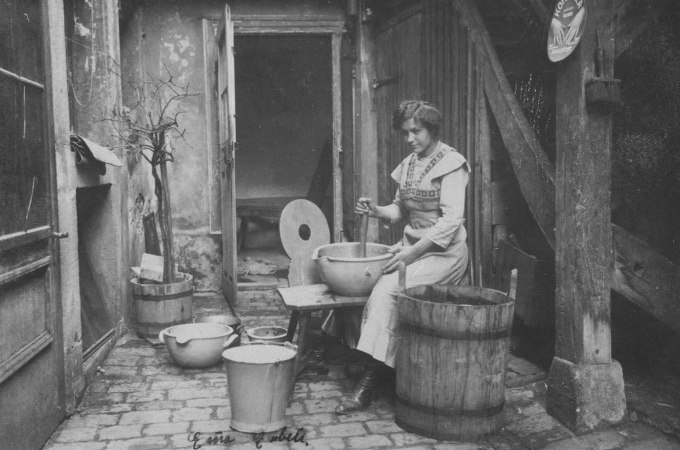 Eine Frau sitzt in einem Innenhof und w&auml;scht Kleidung in einer Sch&uuml;ssel. Um sie herum stehen Eimer und ein Waschbrett. Die Szene wirkt historisch., &copy; Stadt N&uuml;rtingen