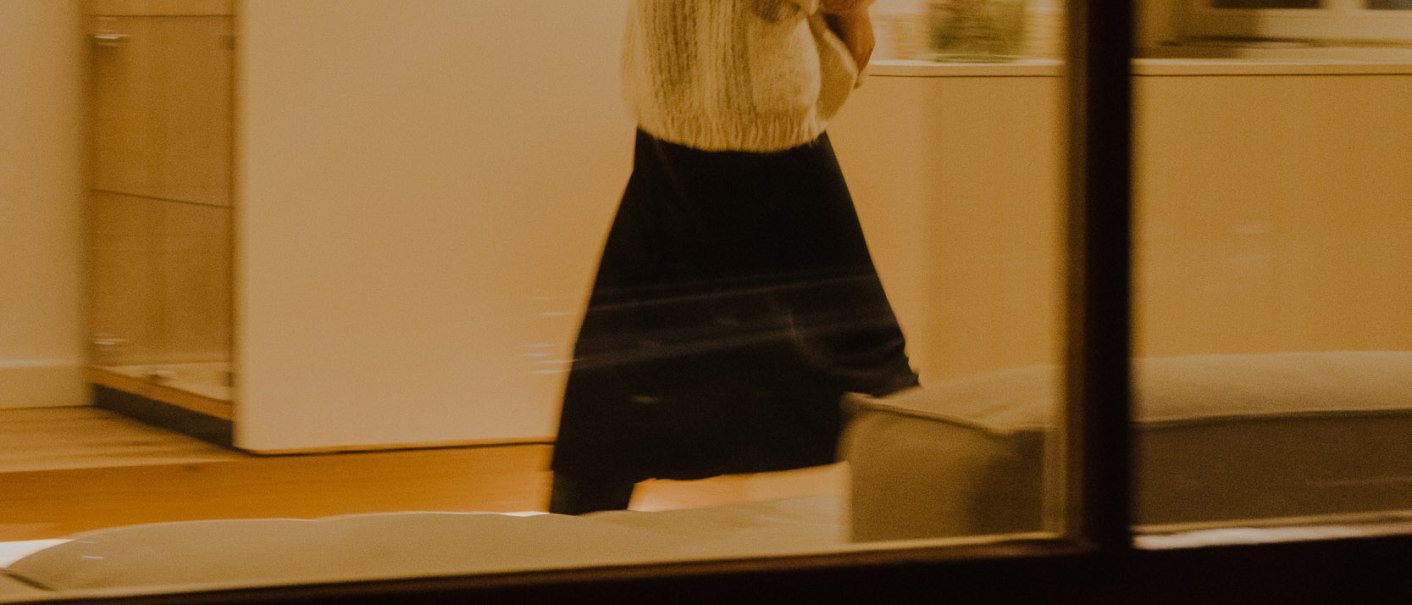 Eine Frau in einem hellen Raum, durch ein Fenster fotografiert. Sie trägt einen weißen Pullover und einen langen schwarzen Rock. Die Beleuchtung ist warm., © Peter Oliver Wolff / DAS EDITH