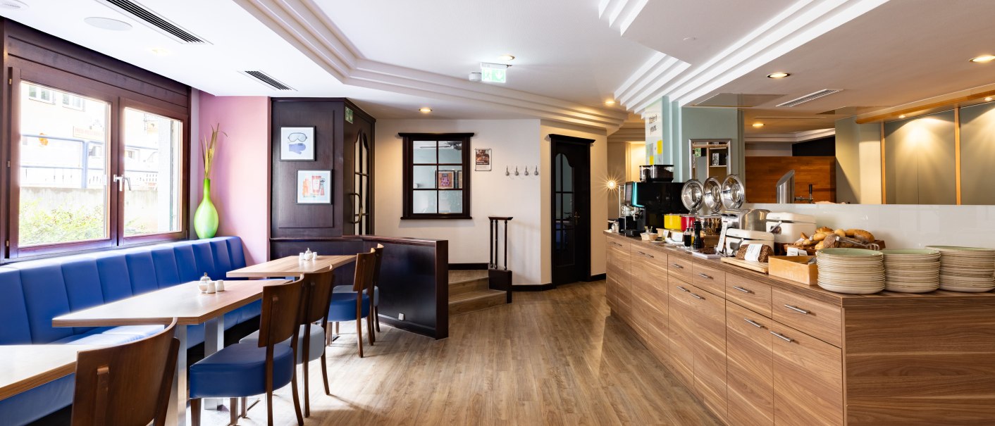 Ein heller Fr&uuml;hst&uuml;cksraum mit blauen Sitzb&auml;nken, Holzboden und einem Buffet mit Geschirr und Speisen. Gro&szlig;e Fenster lassen Tageslicht herein., &copy; Novum  Hospitality