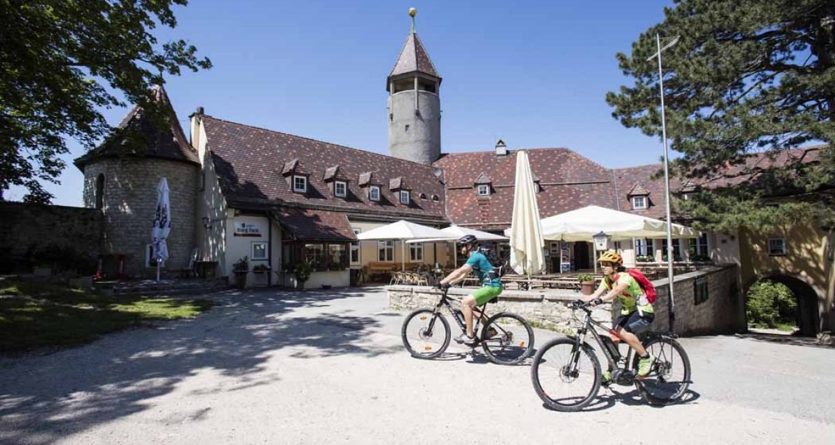 Zwei Radfahrer vor der Burg Teck. Im Hintergrund ein Turm und Sonnenschirme. Der Himmel ist klar und blau., &copy; Schw&auml;bische Alb Tourismusverband e.V.