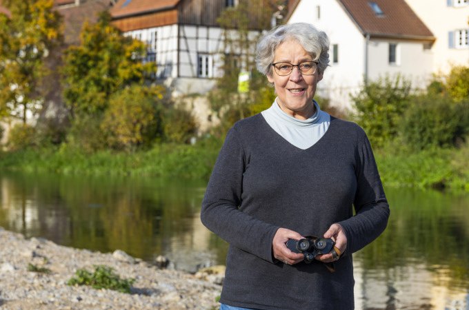 Eine ältere Frau mit Fernglas steht am Flussufer, im Hintergrund Fachwerkhäuser und herbstliche Bäume., © Stadt Besigheim Eine ältere Frau mit Fernglas steht am Flussufer, im Hintergrund Fachwerkhäuser und herbstliche Bäume., © Stadt Besigheim