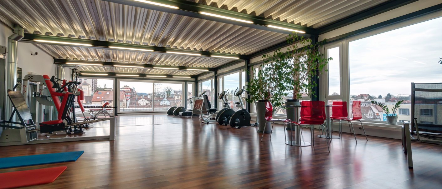 Modernes Fitnessstudio mit Holzboden, Fitnessgeräten und großen Fenstern, die einen Blick auf die Stadt bieten. Rote Stühle und Pflanzen dekorieren den Raum., © City Hotel Reutlingen