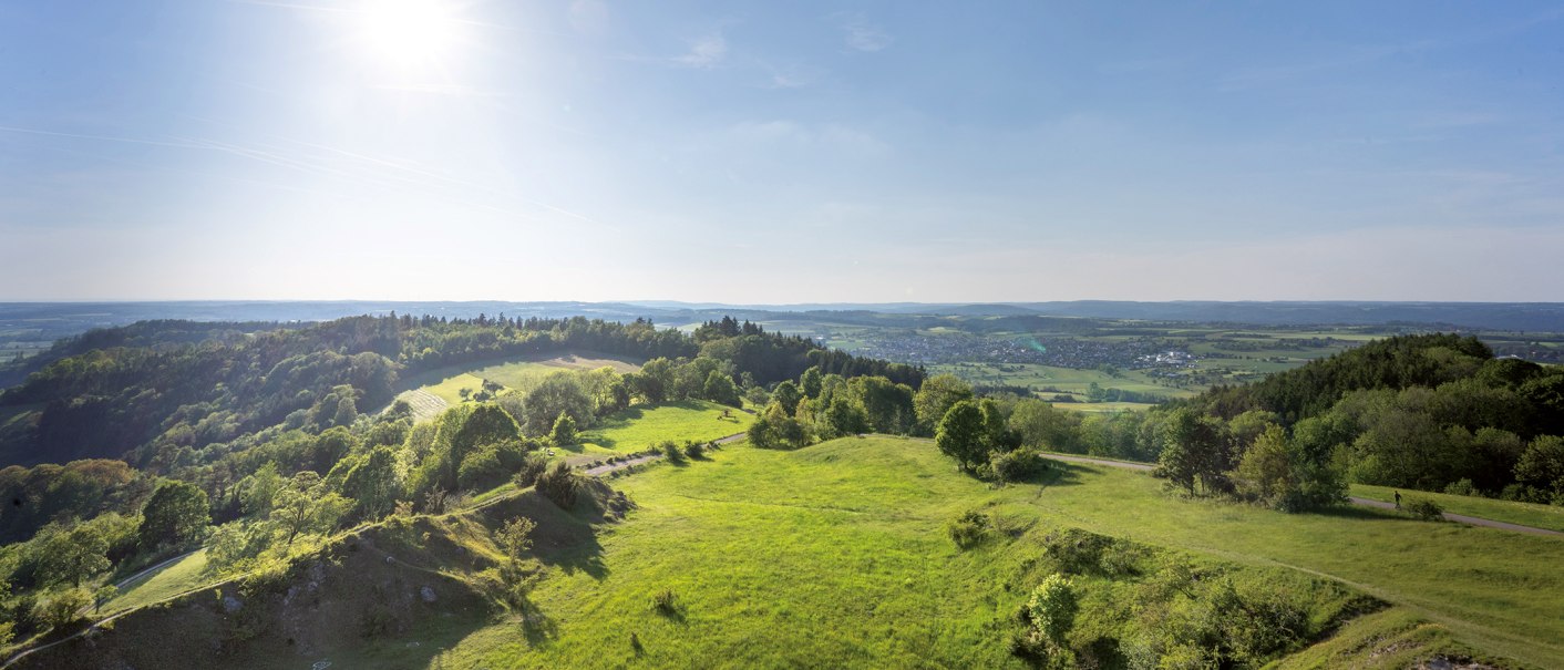 Weite Landschaft mit grünen Hügeln und Wäldern unter klarem Himmel, im Hintergrund eine Stadt. Die Sonne scheint hell., © Stuttgart-Marketing GmbH, Martina Denker