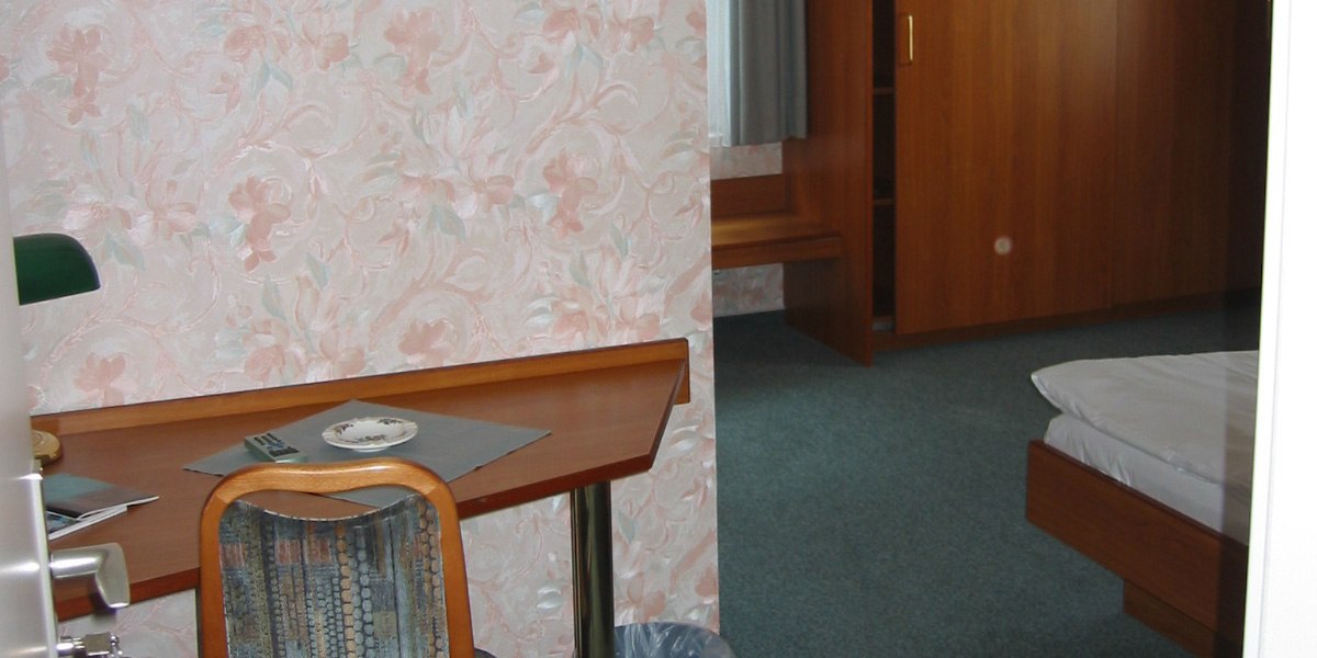 Ein Hotelzimmer mit floral gemusterter Tapete, Schreibtisch, Stuhl, Bett und Holzschrank., © Hotel Wirt am Berg