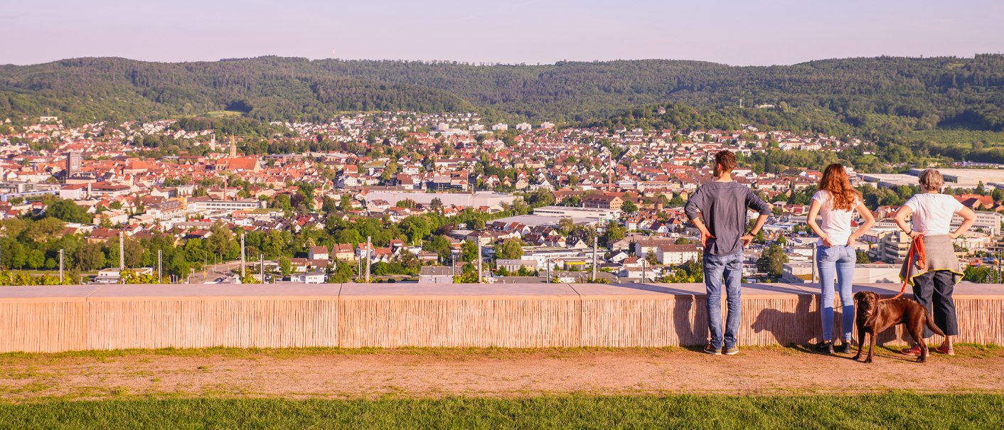 Drei Personen und ein Hund stehen auf einer Aussichtsplattform und blicken auf eine Stadt mit bewaldeten H&uuml;geln im Hintergrund., &copy; Stadt Schorndorf