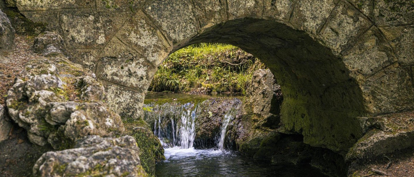 Ein kleiner Wasserfall flie&szlig;t unter einer alten Steinbr&uuml;cke hindurch, umgeben von Felsen und gr&uuml;ner Vegetation., &copy; Stuttgart-Marketing GmbH, Sarah Schmid